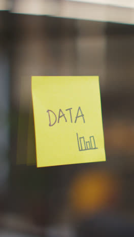 Vertical-Video-Close-Up-Of-Woman-Putting-Sticky-Note-With-Data-Written-On-It-Onto-Transparent-Screen-In-Office